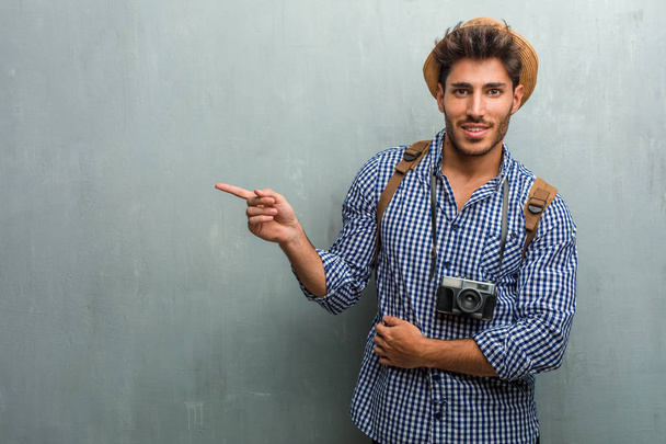Молодой красивый путешественник в соломенной шляпе, рюкзаке и фотоаппарате, указывая в сторону, улыбаясь удивлен, представляя что-то, естественное и случайное
 - Фото, изображение
