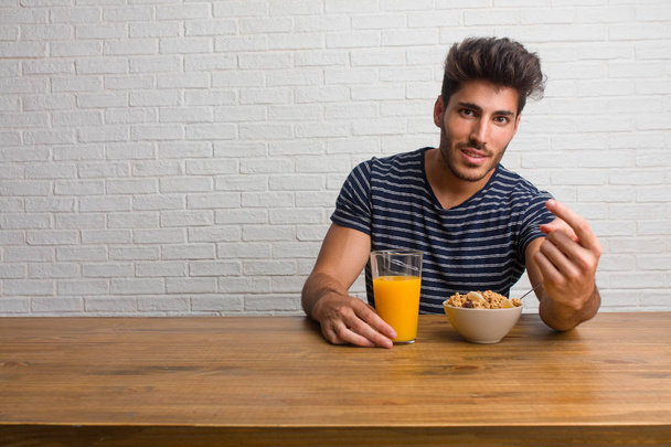 Young handsome and natural man sitting on a table inviting to come, confident and smiling making a gesture with hand, being positive and friendly. Having a breakfast, includes orange juice and a cereals bowl. - 写真・画像