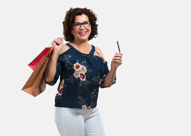 Middle aged woman cheerful and smiling, very excited holding the new bank card and shopping bags, ready to go shopping - Photo, Image