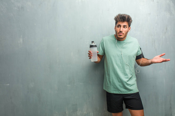 Człowiek młody fitness z butelka sportowa ścianę grunge, wątpienie i wzruszając ramionami, noszenie opaski z telefonu. - Zdjęcie, obraz