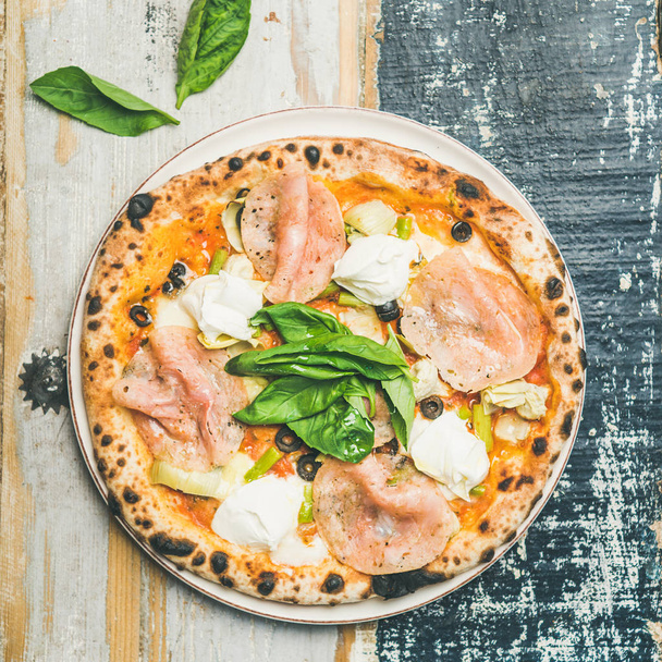 Freshly baked pizza with artichokes, smoked turkey ham, olives, cream cheese and green basil leaves over rustic wooden background - Foto, afbeelding