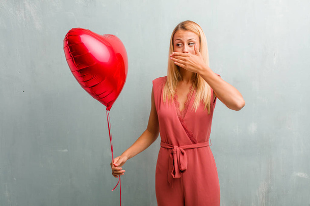 Portrait of young elegant blonde woman covering mouth, symbol of silence and repression, trying not to say anything. Holding a red heart balloon. - Photo, Image