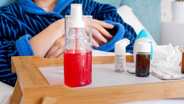 Closeup photo of throat spray, glass of water and pills on wooden tray in bed of sick woman - 写真・画像