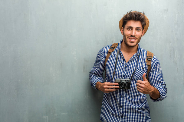 Young handsome traveler man wearing a straw hat, a backpack and a photo camera cheerful and excited, smiling and raising her thumb up, concept of success and approval, ok gesture - Foto, Imagen