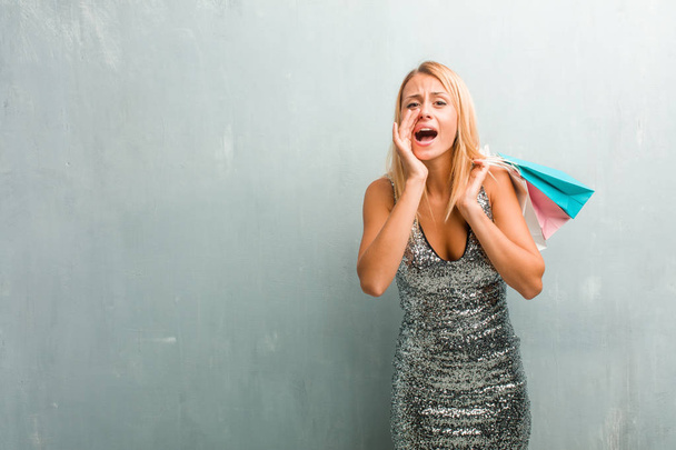 Portret van jonge elegante blonde vrouw schreeuwen gelukkig, verrast door een aanbieding of promotie, gapende, springen en trots. Holding boodschappentas. - Foto, afbeelding