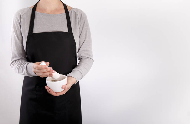 Woman chef grinding herbs in mortar wearing black apron gray background copy space - Photo, Image