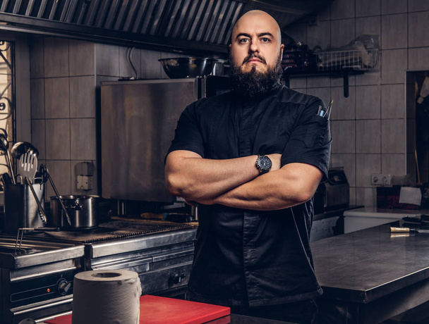 Mutfakta çapraz kolları olan siyah üniforma ayakta acımasız sakallı Şef aşçı. - Fotoğraf, Görsel