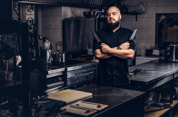 Brodaty kucharz w czarnym mundurze posiada nóż i stojąc z rękami skrzyżowanymi w kuchni. - Zdjęcie, obraz