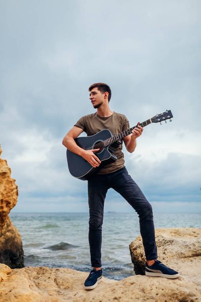 Jeune homme avec guitare acoustique jouant seul sur la plage entourée de rochers le jour de la pluie
 - Photo, image