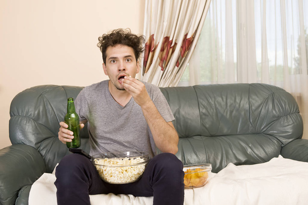 Joven emocionado con el control remoto y palomitas de maíz en el sofá viendo la televisión
 - Foto, imagen