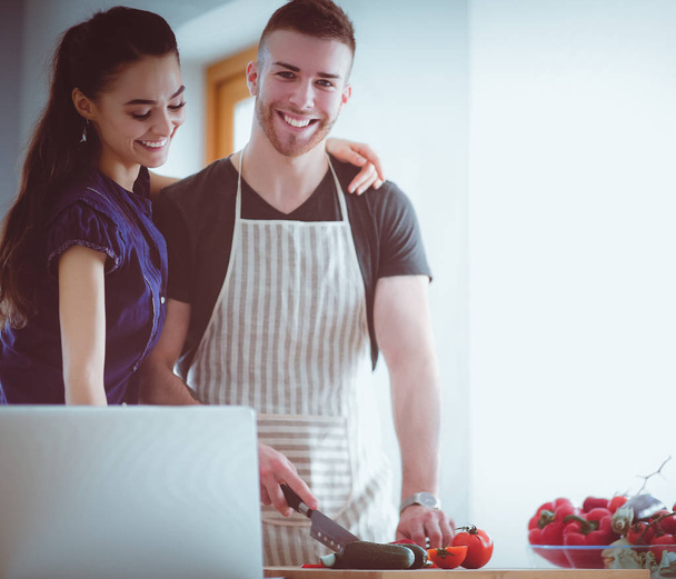 Young man cutting vegetables and woman standing with laptop in the kitchen - Fotoğraf, Görsel