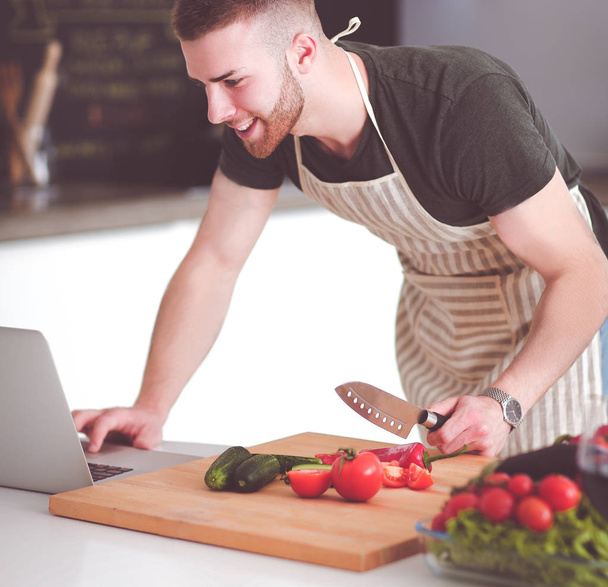 Portrait of man cooking vegetable in the kitchen while looking at a laptop computer on the table - 写真・画像