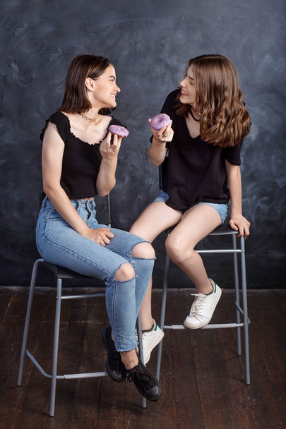Pretty happy teenage girls with donuts  siting on chairs and having fun. Portrait of joyful smiling girls with donuts on black background. Good mood, diet concept. - Fotó, kép