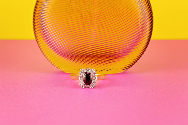 Garnet solitaire ring surrounded by diamonds in a square shape with gold band in front of a transparent perfume bottle isolated on a pink surface with a yellow background - Photo, Image