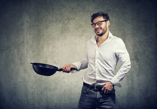 Young casual man holding frying pan and looking excited happy with cooking food on gray background - Photo, image