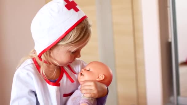 little girl is playing doctor. A child in a doctors suit soothes a toy doll - Materiał filmowy, wideo