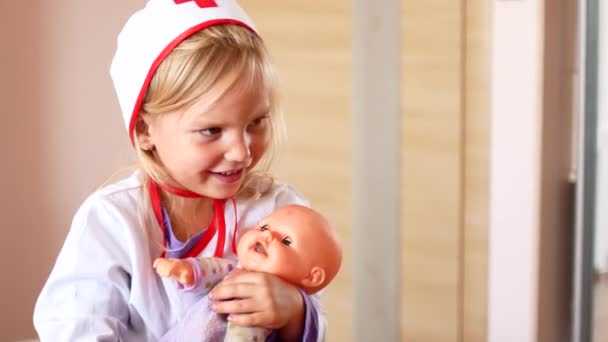 little girl is playing doctor. A child in a doctors suit soothes a toy doll - Materiał filmowy, wideo