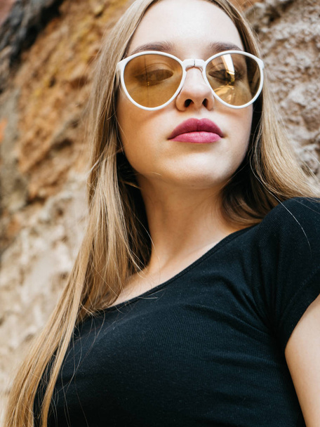 Portrait of trendy casual young woman in black wear and sunglasses with long blond hair posing near urban wall along  - Valokuva, kuva