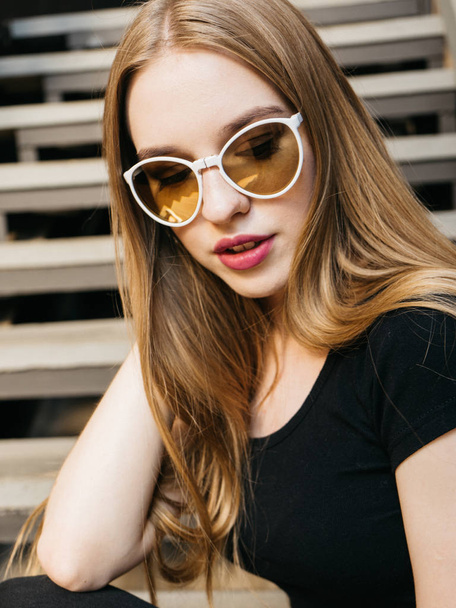 Portrait of trendy casual young woman in black wear and sunglasses with long blond hair posing near urban wall along  - 写真・画像
