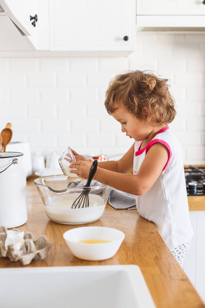 Little girl preparing dough for pancakes at the kitchen. Concept of food preparation, white kitchen on background. Casual lifestyle photo series in real life interior - Foto, imagen