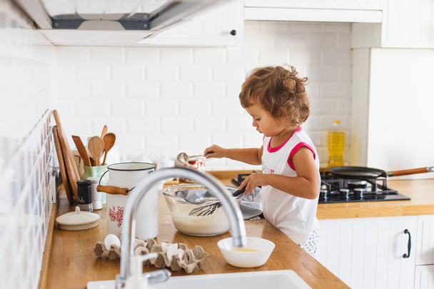 Little girl preparing dough for pancakes at the kitchen. Concept of food preparation, white kitchen on background. Casual lifestyle photo series in real life interior - Foto, immagini