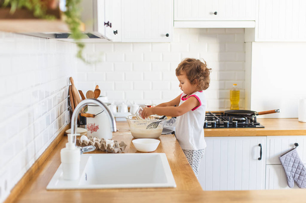 Little girl preparing dough for pancakes at the kitchen. Concept of food preparation, white kitchen on background. Casual lifestyle photo series in real life interior - Valokuva, kuva