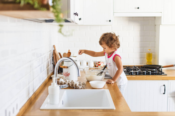 Little girl preparing dough for pancakes at the kitchen. Concept of food preparation, white kitchen on background. Casual lifestyle photo series in real life interior - Foto, afbeelding