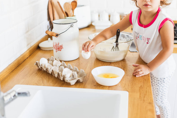 Little girl preparing dough for pancakes at the kitchen. Concept of food preparation, white kitchen on background. Casual lifestyle photo series in real life interior - Photo, image