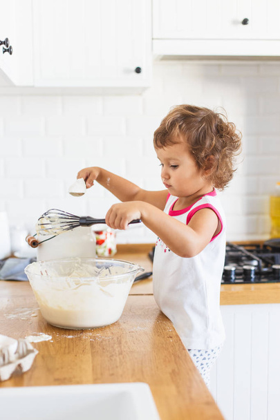Little girl preparing dough for pancakes at the kitchen. Concept of food preparation, white kitchen on background. Casual lifestyle photo series in real life interior - Photo, Image