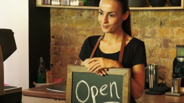 Un trabajador de una cafetería sostiene un letrero abierto y mira al cliente
. - Imágenes, Vídeo