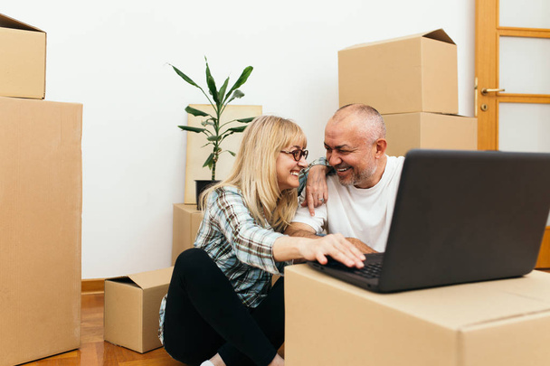 Middle aged couple sitting between boxes in their new apartment and using a laptop for some interior decorating ideas. Real estate theme. - Foto, immagini