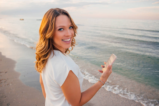 Young beautiful smiling blonde hair curvy model posing on the beach with smartphone in her hand wearing white dress - oversize model with long curly hair on seaside looking into camera while texting - Photo, image