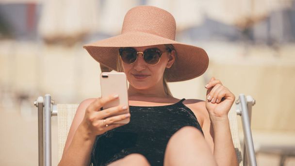 Young beautiful blonde hair curvy woman lying on the beach with smartphone, wearing black chenille swimsuit and pink hat - Фото, изображение