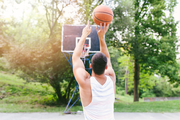 Young athletic man playing basketball in a beautiful park surrounded with trees - Фото, изображение