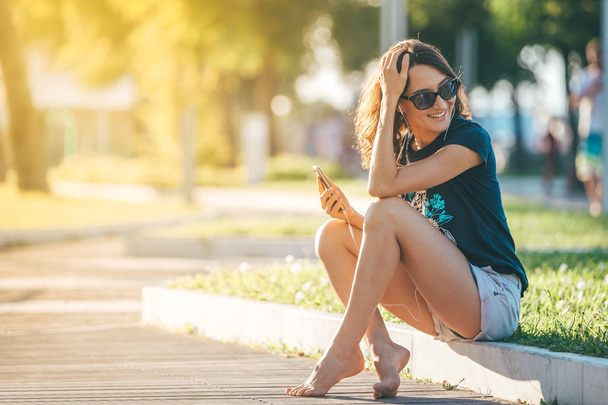 Young beautiful happy woman sitting on the lawn with smartphone and headphones listening to music or audio book - talking or chatting on mobile phone - warm autumn or summer sunset image - Фото, зображення