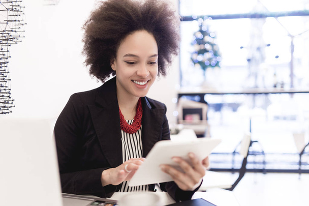 Mujer de negocios afroamericana linda confiada y sonriente sentada en una cafetería moderna y usando su computadora portátil o tableta
. - Foto, imagen