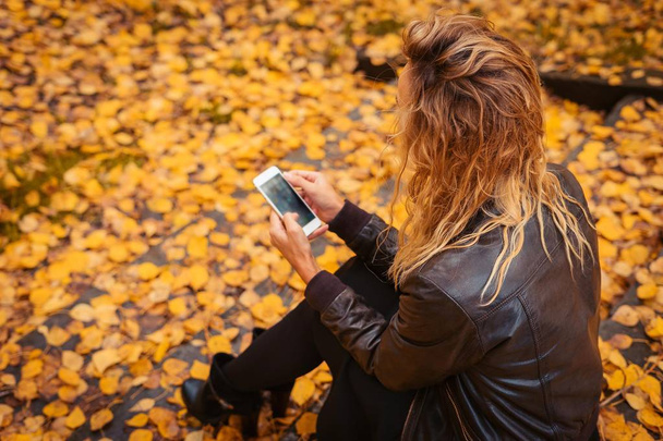 The girl is sitting on the stairs in the autumn forest. Brunette in a leather jacket with a phone in his hands, sms and internet surfing. Yellow leaf fall. - Foto, immagini