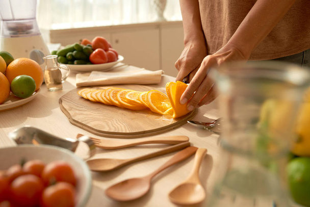 Onherkenbaar vrouw stond op de keukentafel en snijden sappige sinaasappel in plakjes tijdens het koken van lekkere en gezonde snack  - Foto, afbeelding