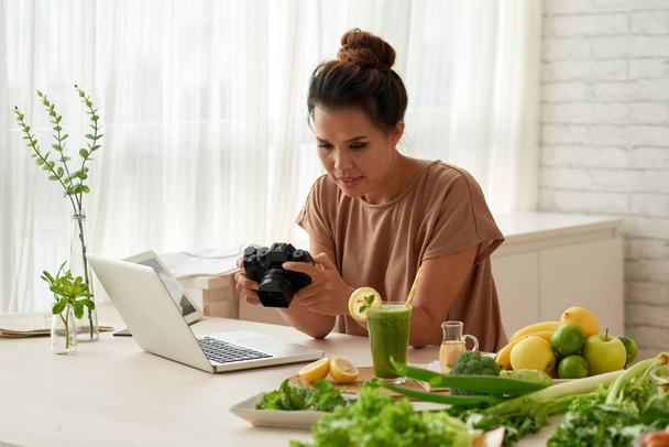 симпатичная азиатская кулинарный блогер, глядя на фотографии на современной камере во время работы над своим блогом дома
 - Фото, изображение