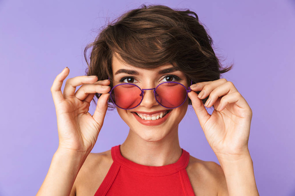 Image of cheerful woman 20s in casual wear and sunglasses looking at you with smile isolated over violet background - Foto, Imagen