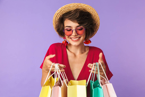 Foto de mulher shopaholic 20s em chapéu de palha segurando sacos de compras de papel colorido com compras isoladas sobre fundo violeta
 - Foto, Imagem