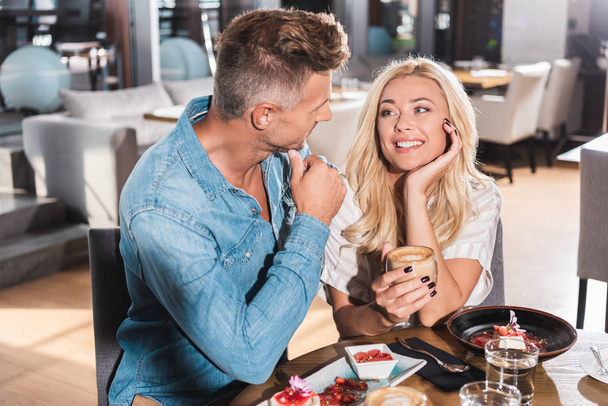 beautiful blonde girlfriend holding cup of coffee and looking at boyfriend at table in cafe  - Foto, Imagen