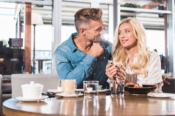 cheerful girlfriend holding cup of coffee and looking at handsome boyfriend at table in cafe  - Foto, afbeelding