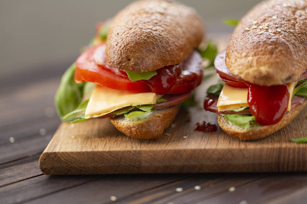 Two baguette sandwiches with salami, cheese, arugula salad, tomatoes and onion on a cutting board. Sandwiches on dark wooden background. Tasty snack. - Foto, immagini