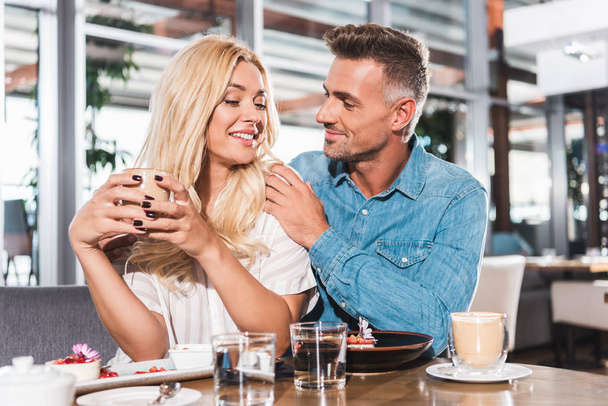 poikaystävä halaus hymyilevä tyttöystävä ja hän tilalla kuppi kahvia pöydässä kahvilassa
 - Valokuva, kuva
