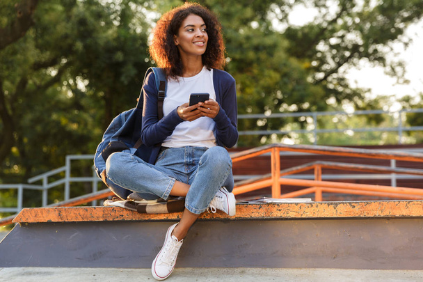 Image of happy african young woman sitting outdoors in park using mobile phone. - Φωτογραφία, εικόνα