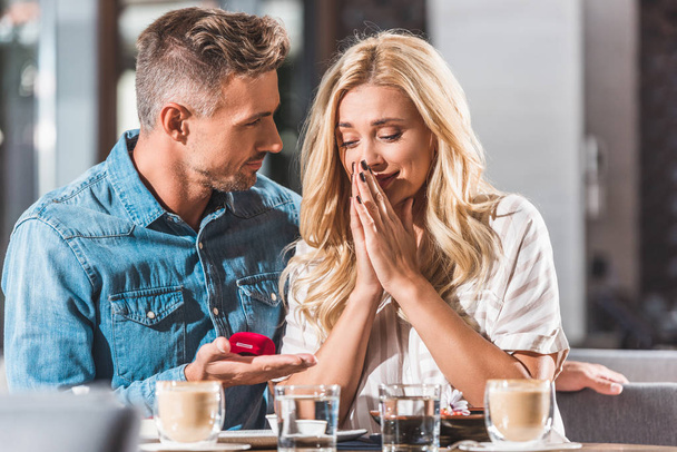 handsome boyfriend making proposal to surprised girlfriend and holding ring box in cafe - Photo, Image