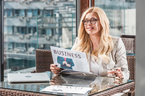 Portret van Glimlachende zakenvrouw met glas water lezen van de krant aan tafel in café - Foto, afbeelding