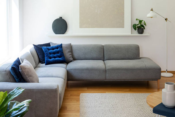 Blue pillows on grey corner sofa in apartment interior with lamp and plant next to poster. Real photo - Photo, Image