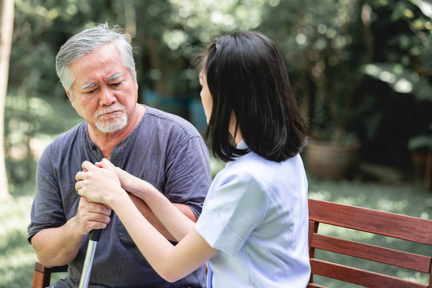 Nurse with patient sitting on bench together talking. Asian old man and young woman sitting together talking. Serious mood. - Photo, Image